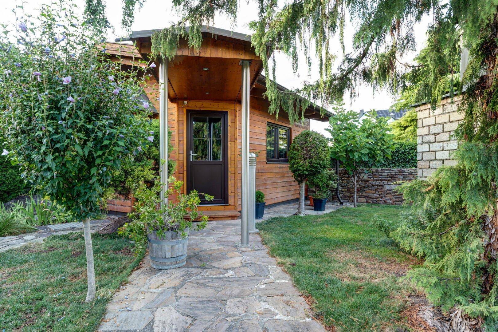 Charmantes Holzhaus im Garten mit Steinweg und grüner Umgebung, umgeben von Bäumen.