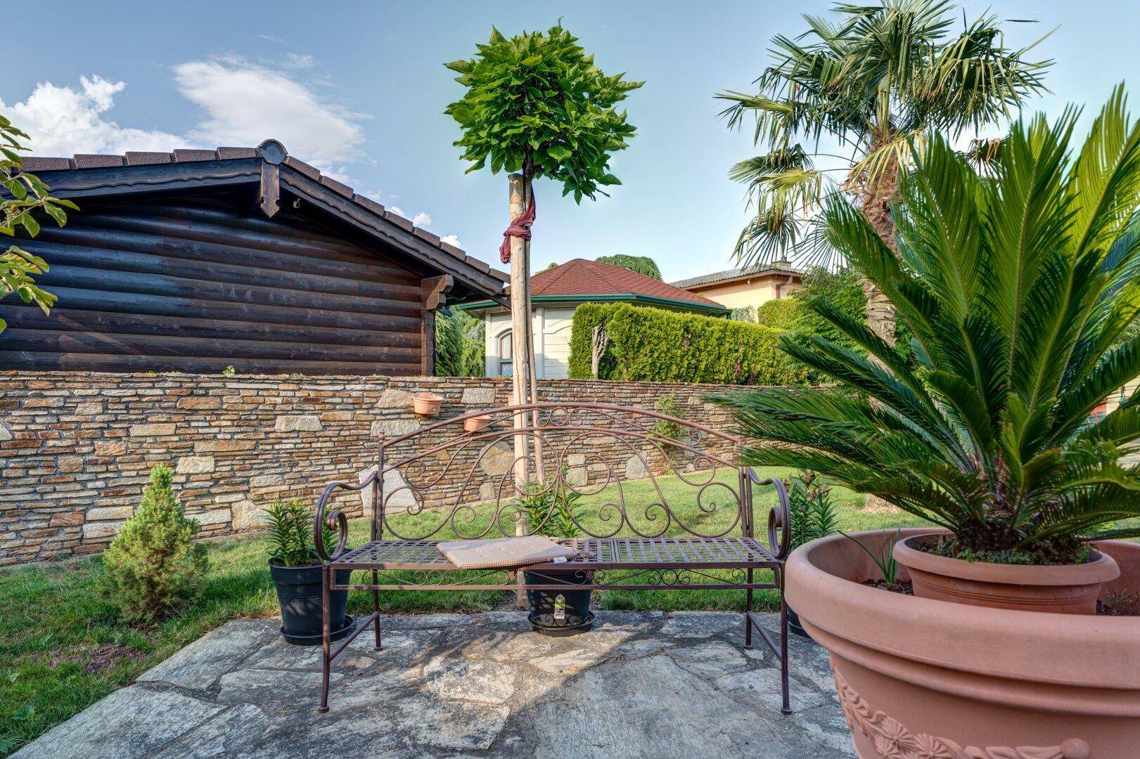 Gemütlicher Sitzbereich im Garten mit Metallbank, Palmen und Steinplatten unter blauem Himmel.