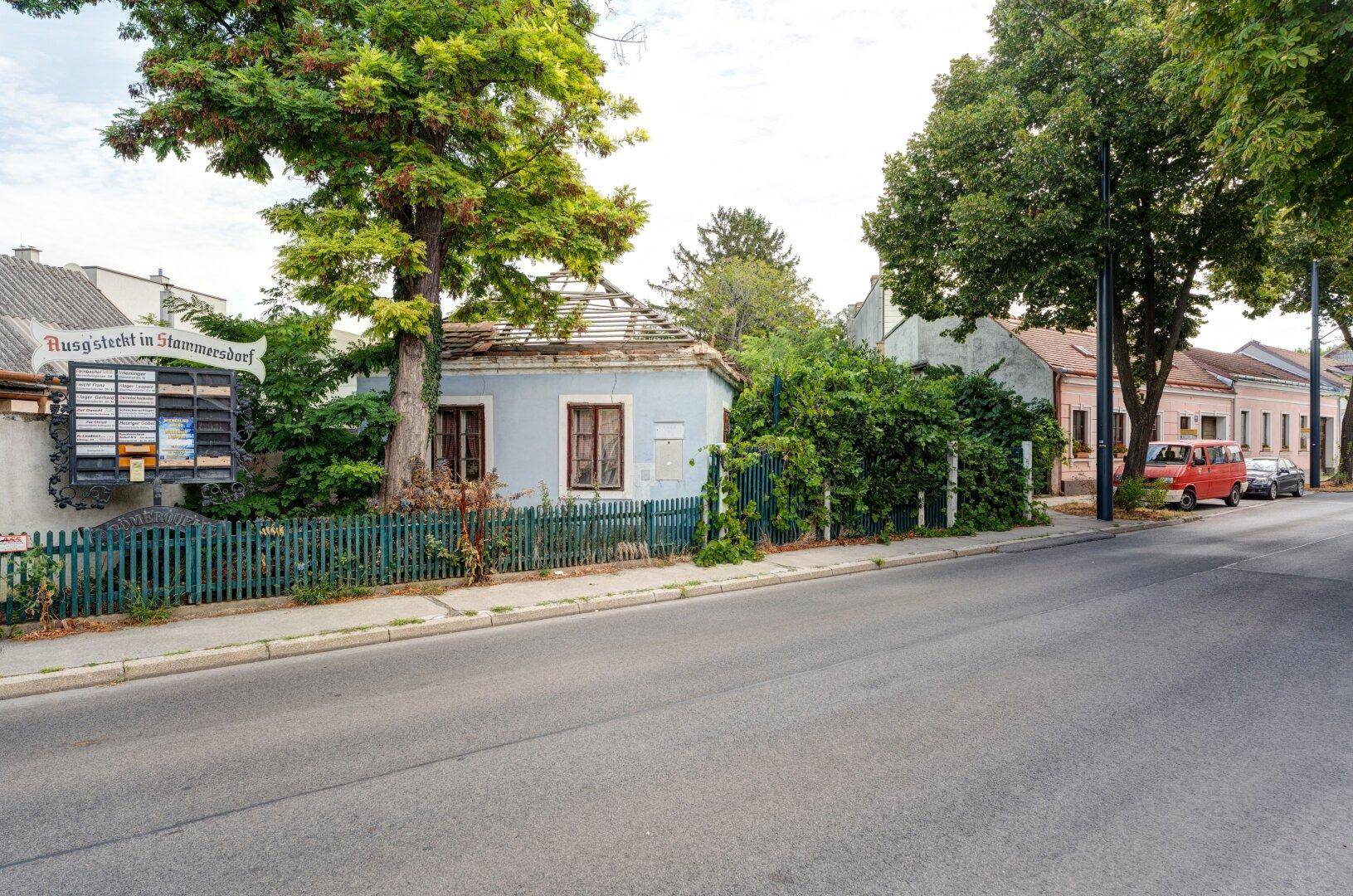 Blaue Fassade eines älteren Gebäudes mit viel Grün, einem Holzzaun und einem Informationsschild.