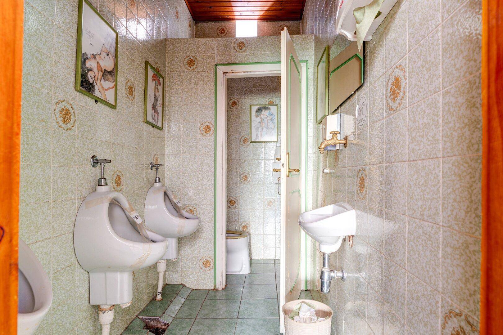 Herren-WC mit mehreren Urinalen, gefliesten Wänden und einem kleinen Waschbecken im Vintage-Stil.