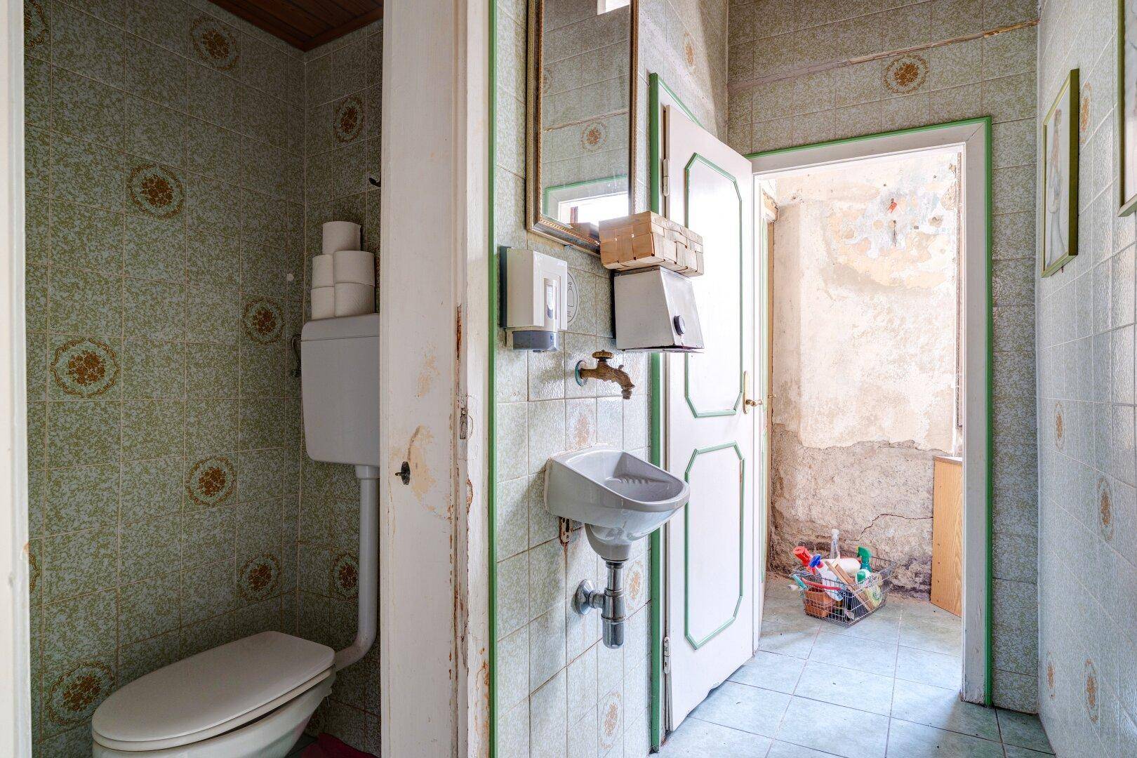 Toilette mit gefliesten Wänden, WC und kleinem Waschbecken, die renovierungsbedürftig erscheint.