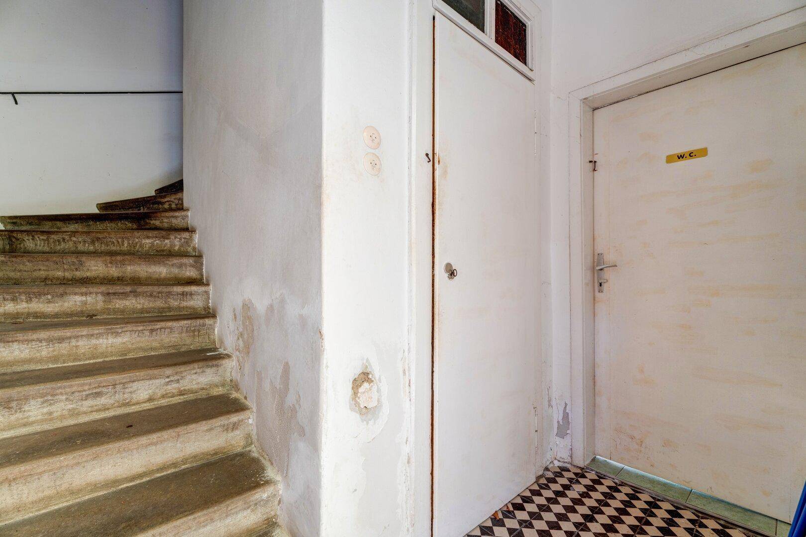 Treppenhaus mit abgenutzten Stufen, schachbrettartigem Fliesenboden und einer WC-Tür.
