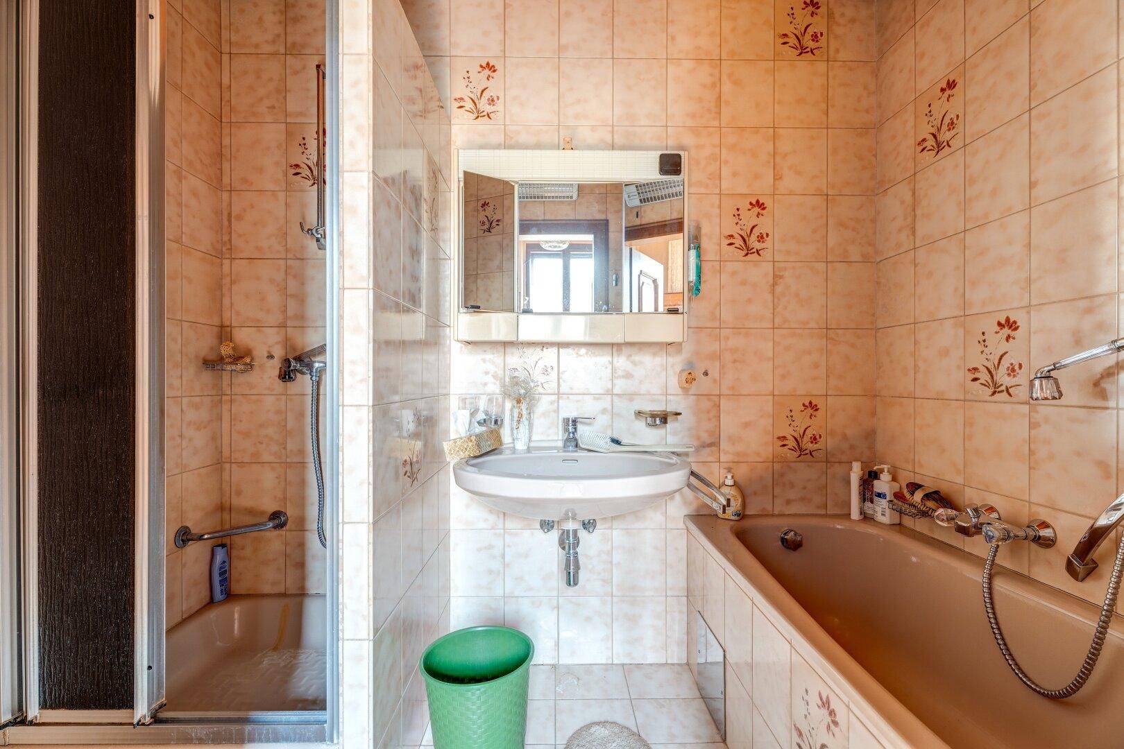 Badezimmer mit beigefarbenen Fliesen, Badewanne und Duschkabine in älterem Zustand.