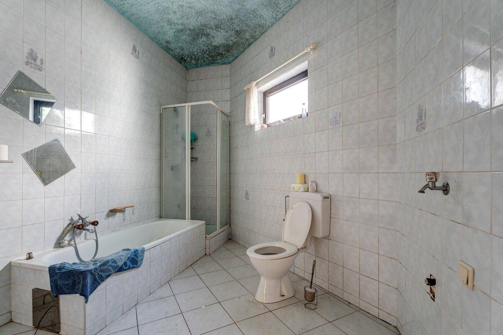 Badezimmer mit weißen Fliesen, Badewanne, Dusche und Toilette in älterem Zustand.
