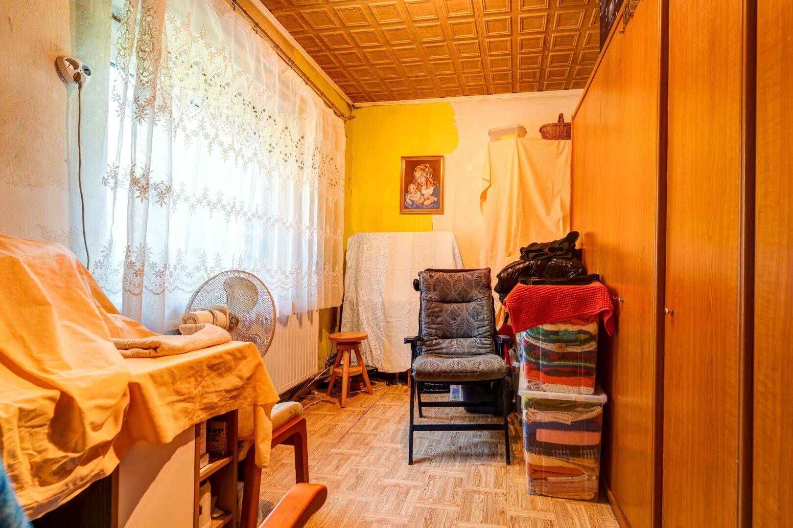 Einfaches Zimmer mit Holzschrank, gemusterter Decke und einem Fenster mit Vorhängen, das als Schlafzimmer dient.