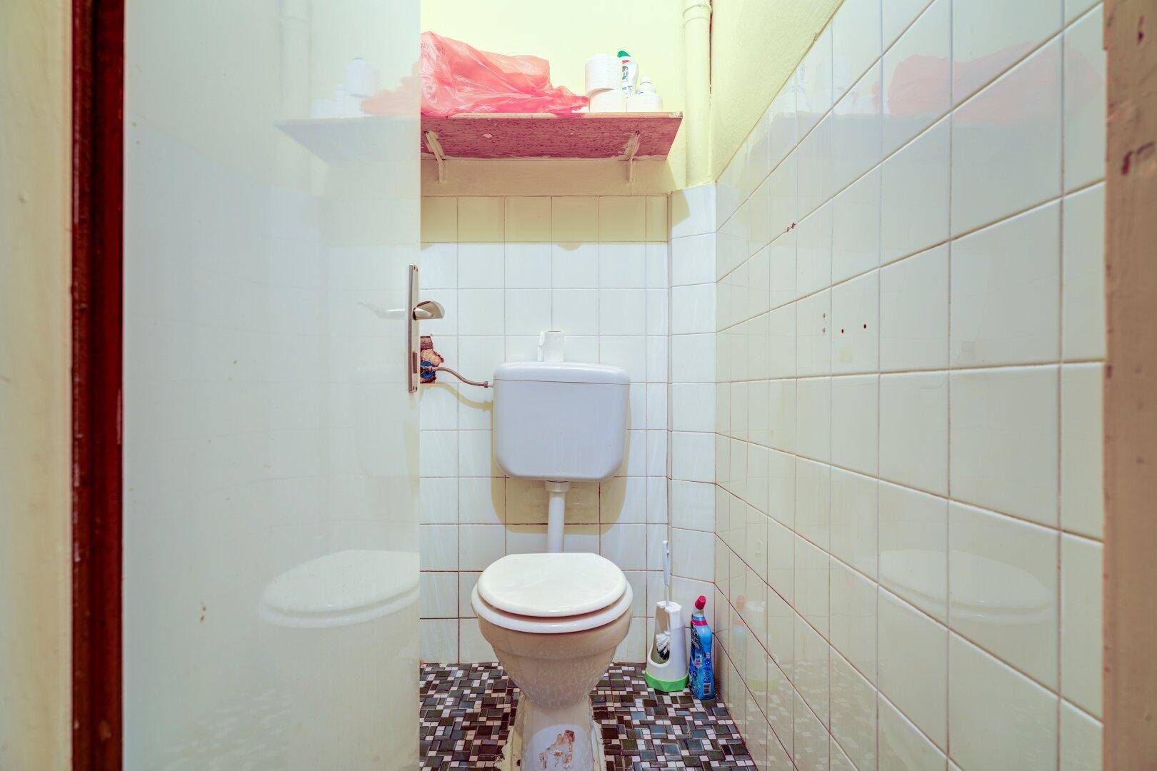 Kleines WC mit weißen Fliesen, dunklem Boden und einer einfachen Toilette in einem schmalen Raum.