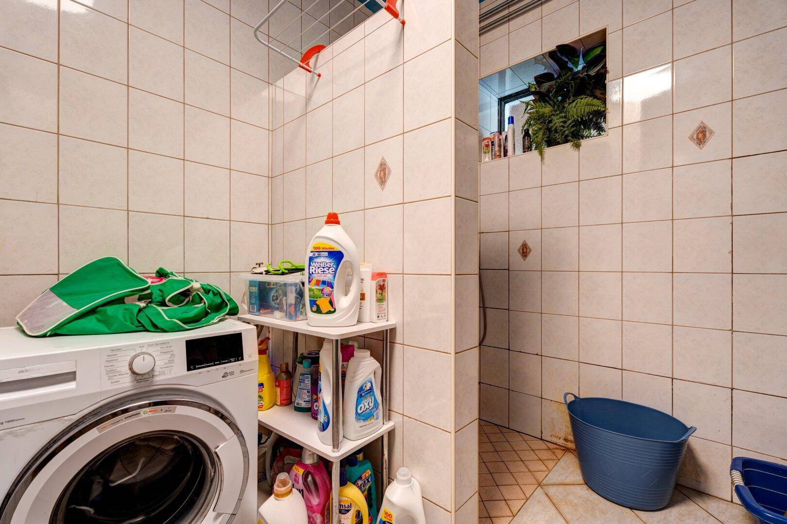 Gefliestes Badezimmer mit Waschmaschine, Duschbereich und praktischen Ablagemöglichkeiten für Utensilien.