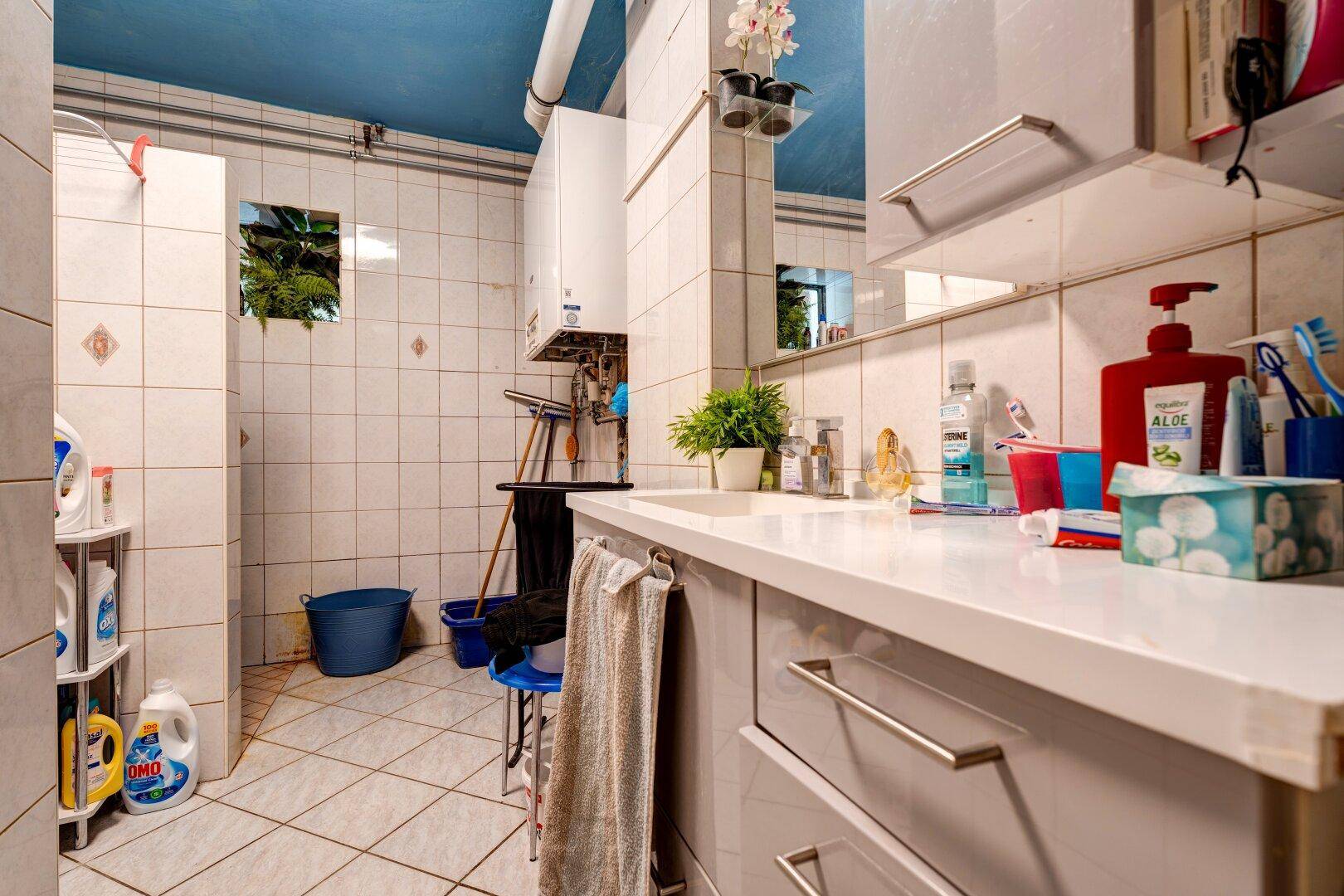 Badezimmer mit Waschtisch, Spiegel, Boiler und gefliesten Wänden in hellem Ambiente, funktional eingerichtet.