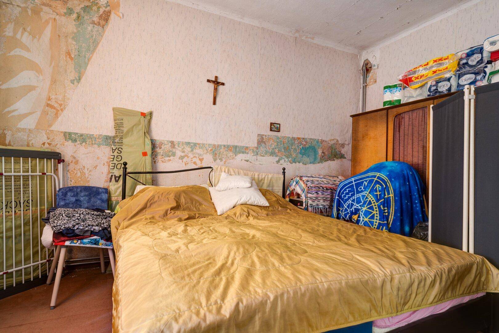 Schlafzimmer mit Doppelbett, abgenutzten Wänden und einem Holzkreuz an der Wand, das einen rustikalen Charme verleiht.
