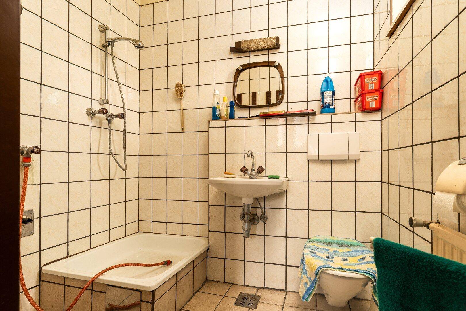 Veraltetes Badezimmer mit weißen Fliesen, kleiner Badewanne und einfacher Ausstattung.