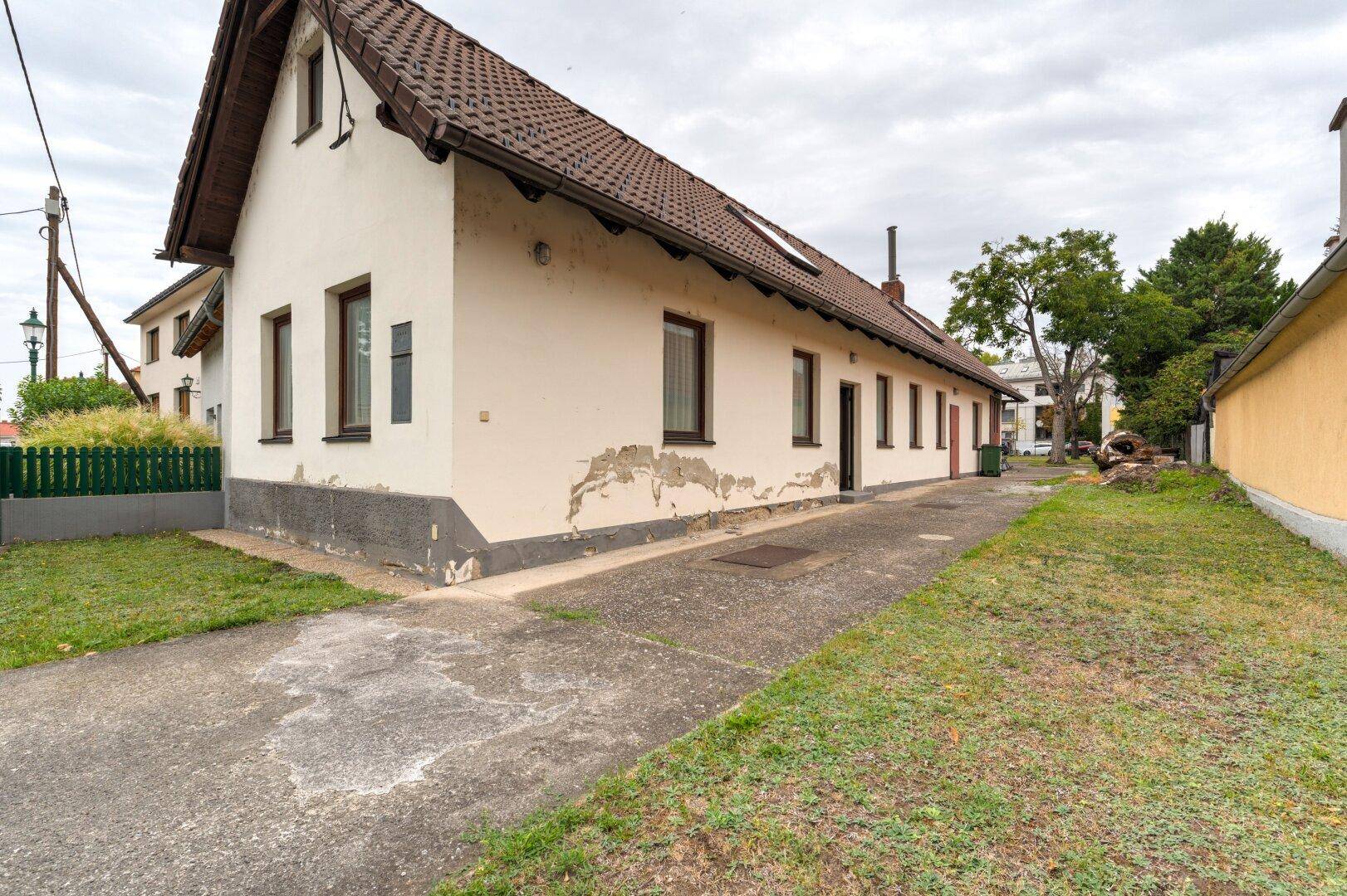 Seitenansicht des Hauses mit bröckelndem Putz, einer Zufahrt und einem kleinen Rasenbereich.