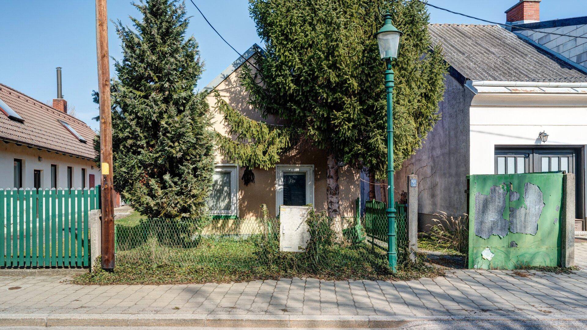 Straßenansicht eines älteren Hauses mit grünem Zaun und altem Baumbestand im Vorgarten.