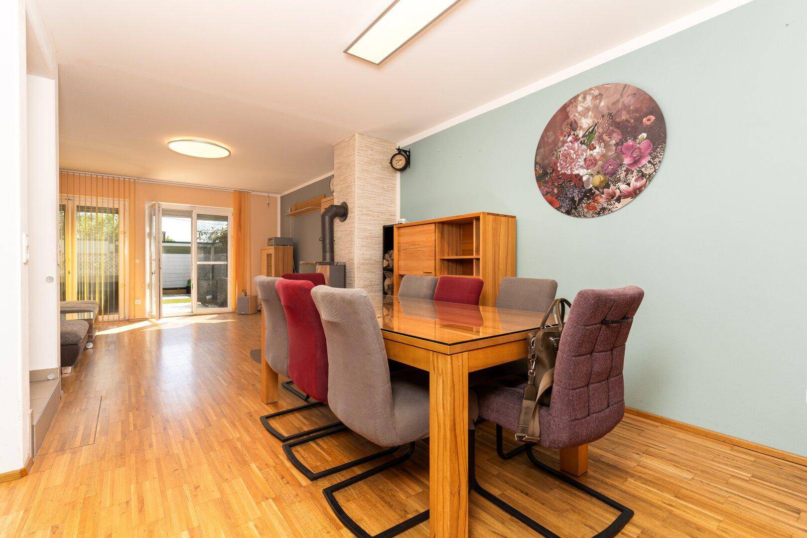 Essbereich mit Holztisch und farbigen Stühlen, offen zum Wohnzimmer und mit Blick auf den Holzofen.