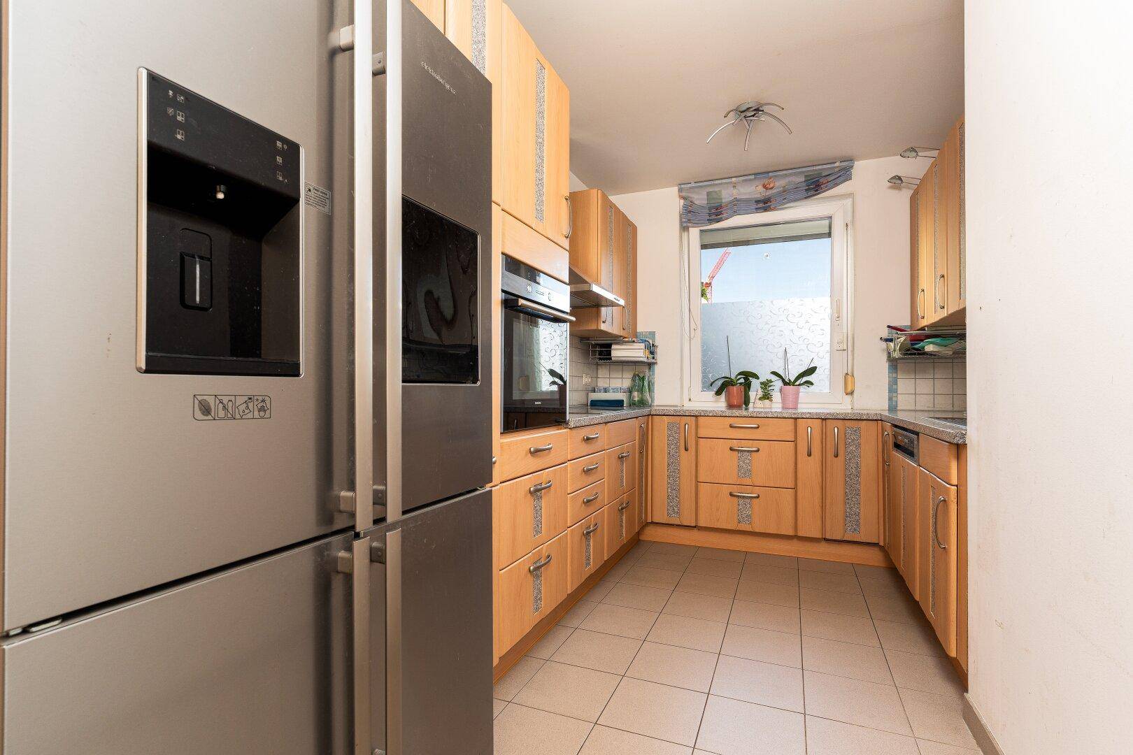 Funktionale Küche mit hellen Holzschränken, Fliesenboden und großem Edelstahlkühlschrank.