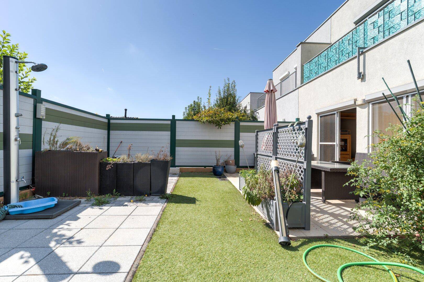Gepflegter Gartenbereich mit Kunstrasen, gepflasterter Terrasse und Außendusche.