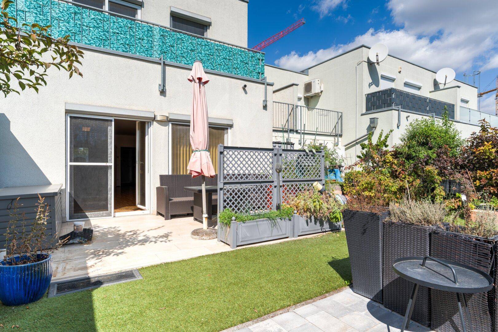 Gartenbereich mit Kunstrasen und Terrasse, ausgestattet mit Gartenmöbeln und Sonnenschirm.