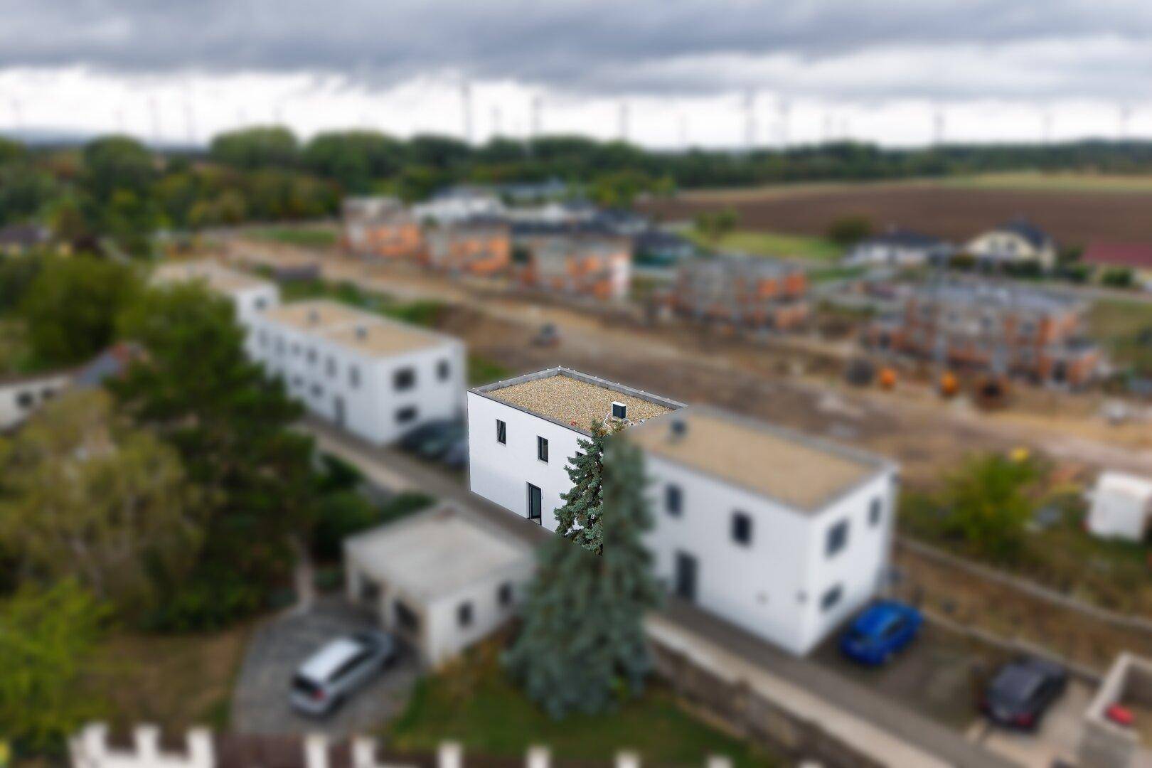 Luftaufnahme einer Neubausiedlung mit mehreren Häusern und Windrädern im Hintergrund.