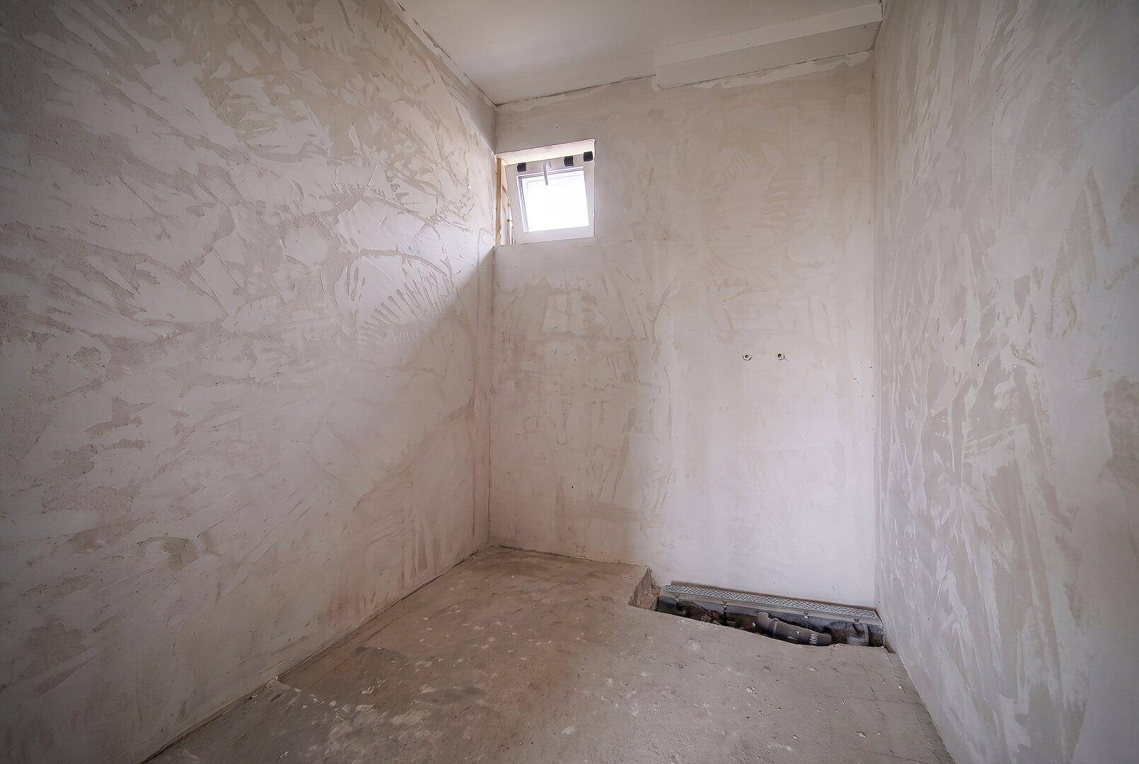 Unfertiges Badezimmer mit Fenster und vorbereiteten Anschlüssen für Sanitäranlagen.