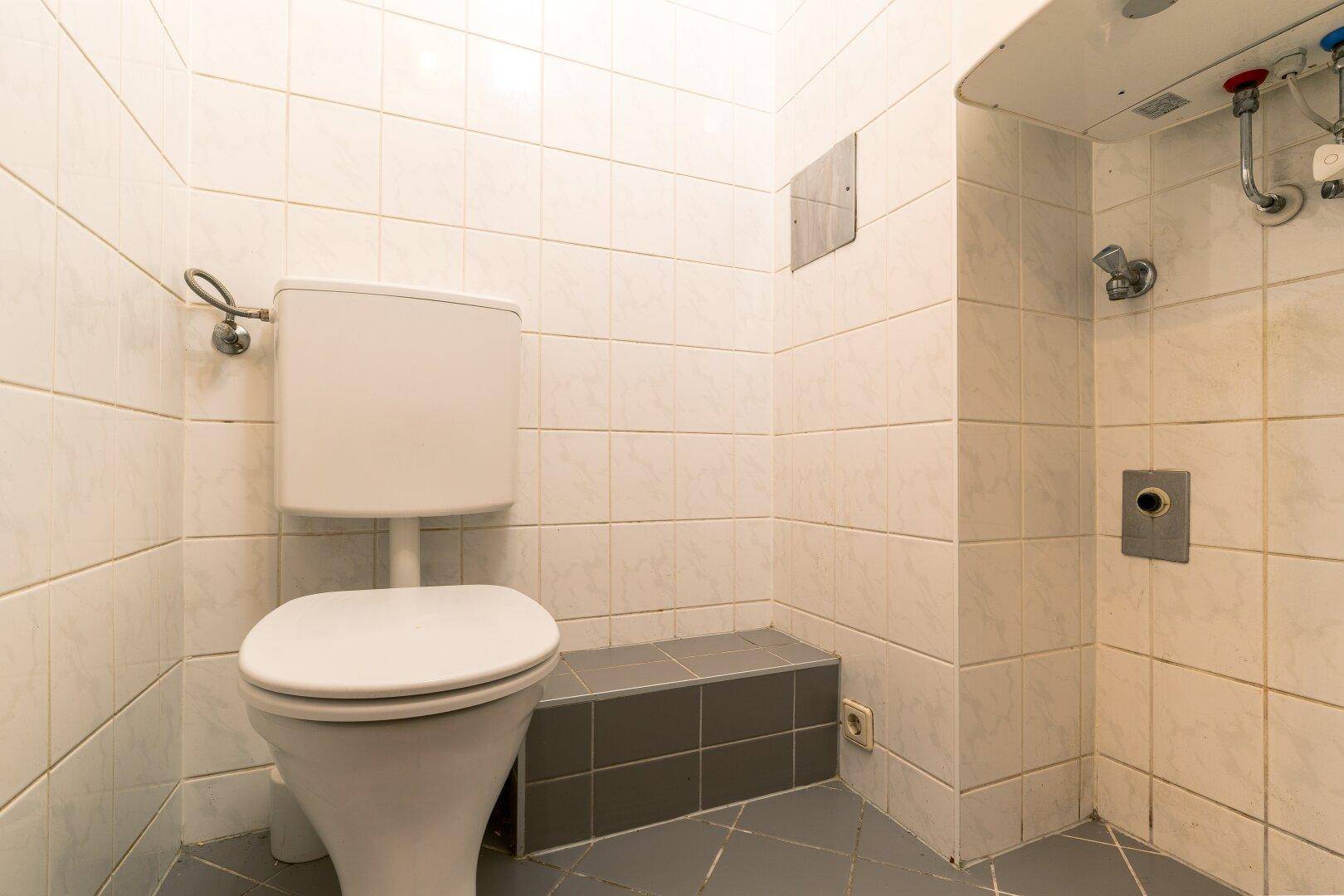 Einfaches WC mit weißen Fliesen an den Wänden und einem grauen Fliesenboden.