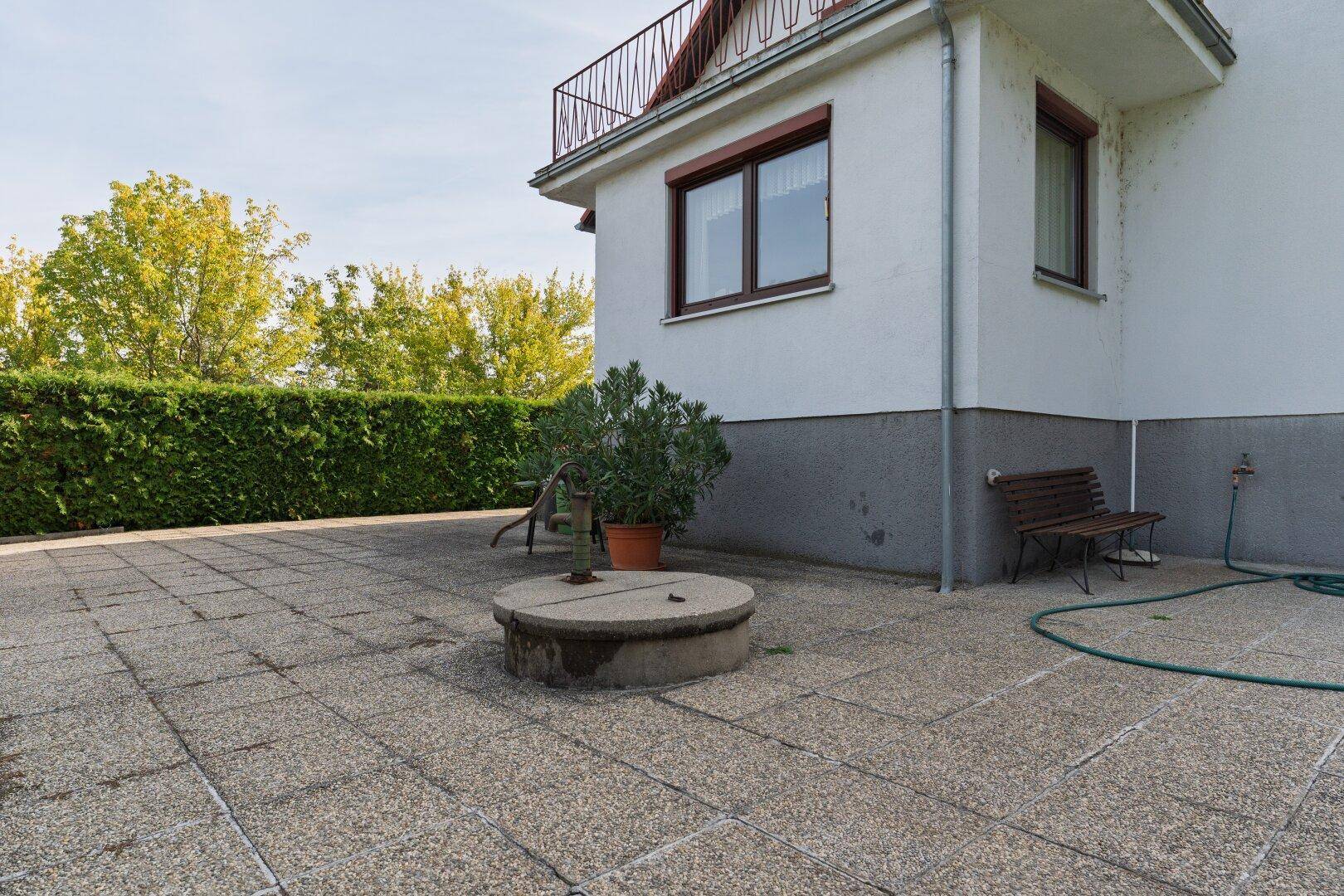 Große gepflasterte Terrasse neben dem Haus mit einem alten Brunnen und einer Hecke im Hintergrund.