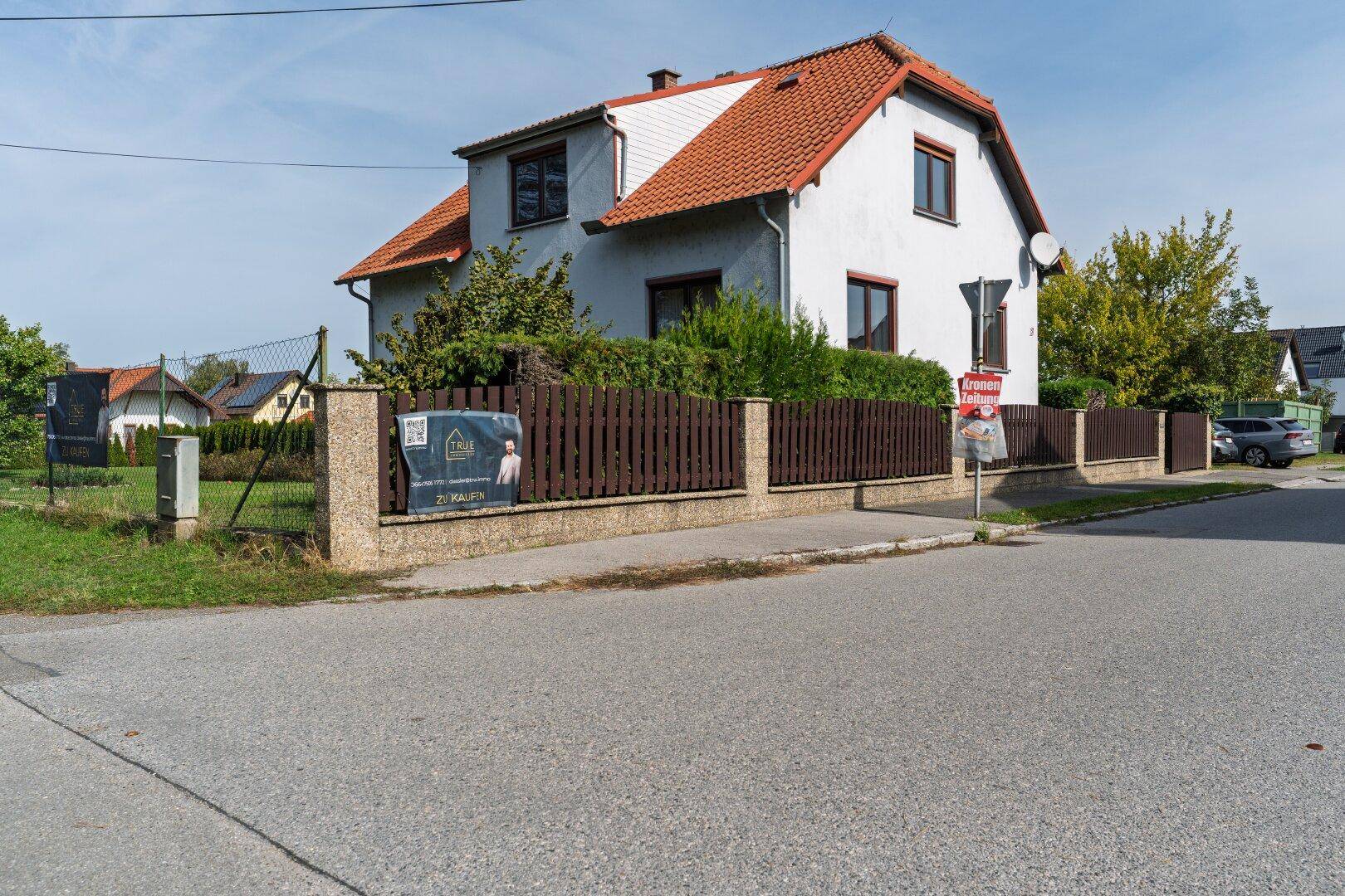 Einfamilienhaus mit rotem Dach und weißer Fassade, umgeben von einem Holzzaun und Bäumen.