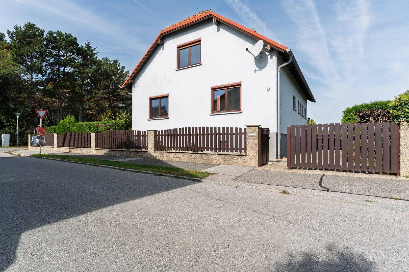 Freistehendes Einfamilienhaus mit Satteldach, weißer Fassade und braunem Holzzaun an der Straße.