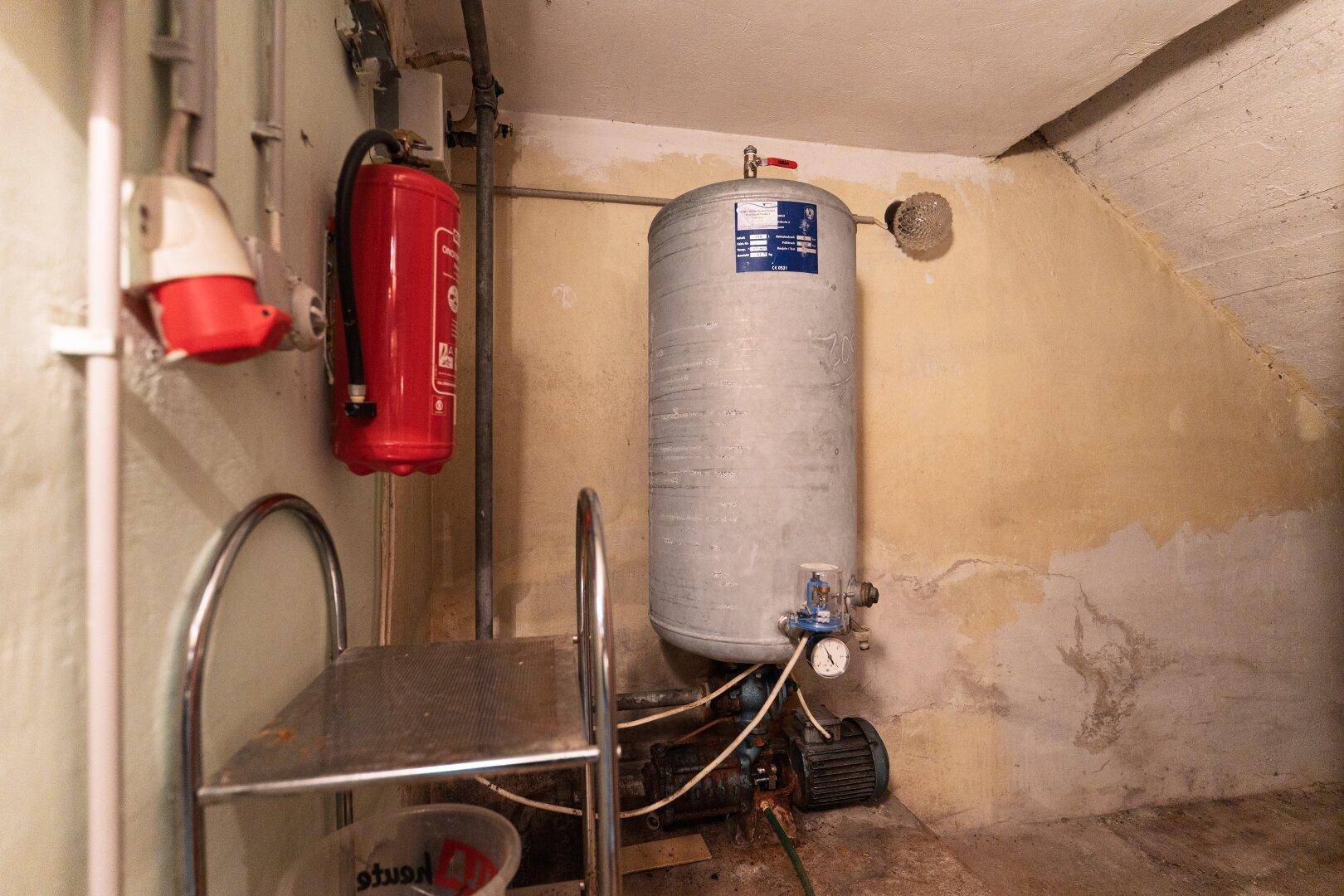 Technischer Raum im Keller mit Warmwasserspeicher, Rohren und einem Feuerlöscher.
