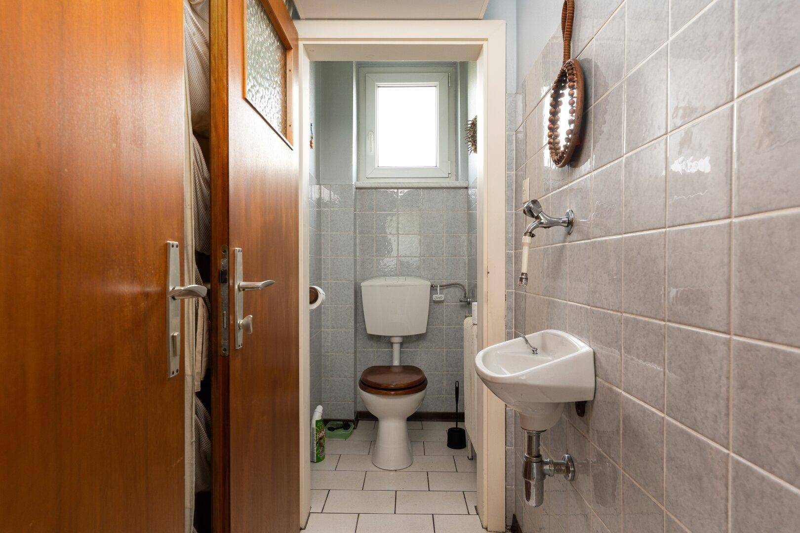 Kleines Gäste-WC mit hellgrauen Fliesen, Fenster und schmalem Waschbecken.