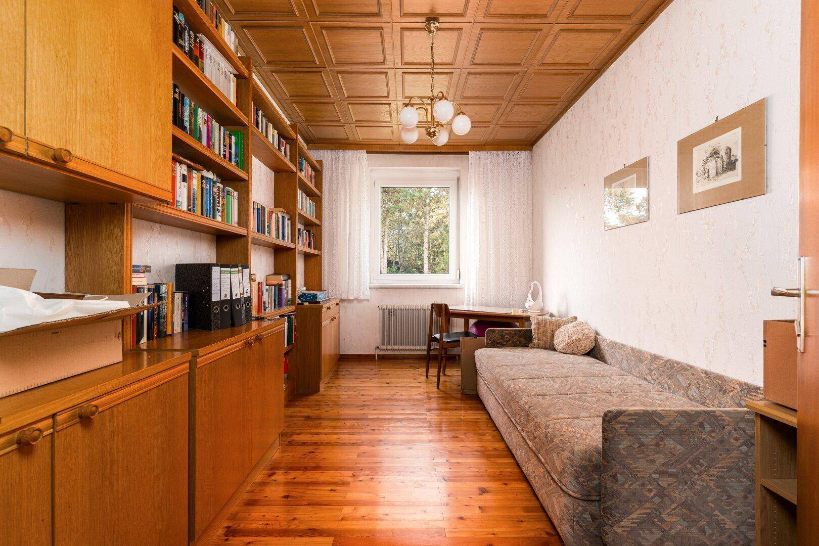 Arbeitszimmer mit Holzdecke, Bücherregalen und einem Sofa, hell durch großes Fenster.