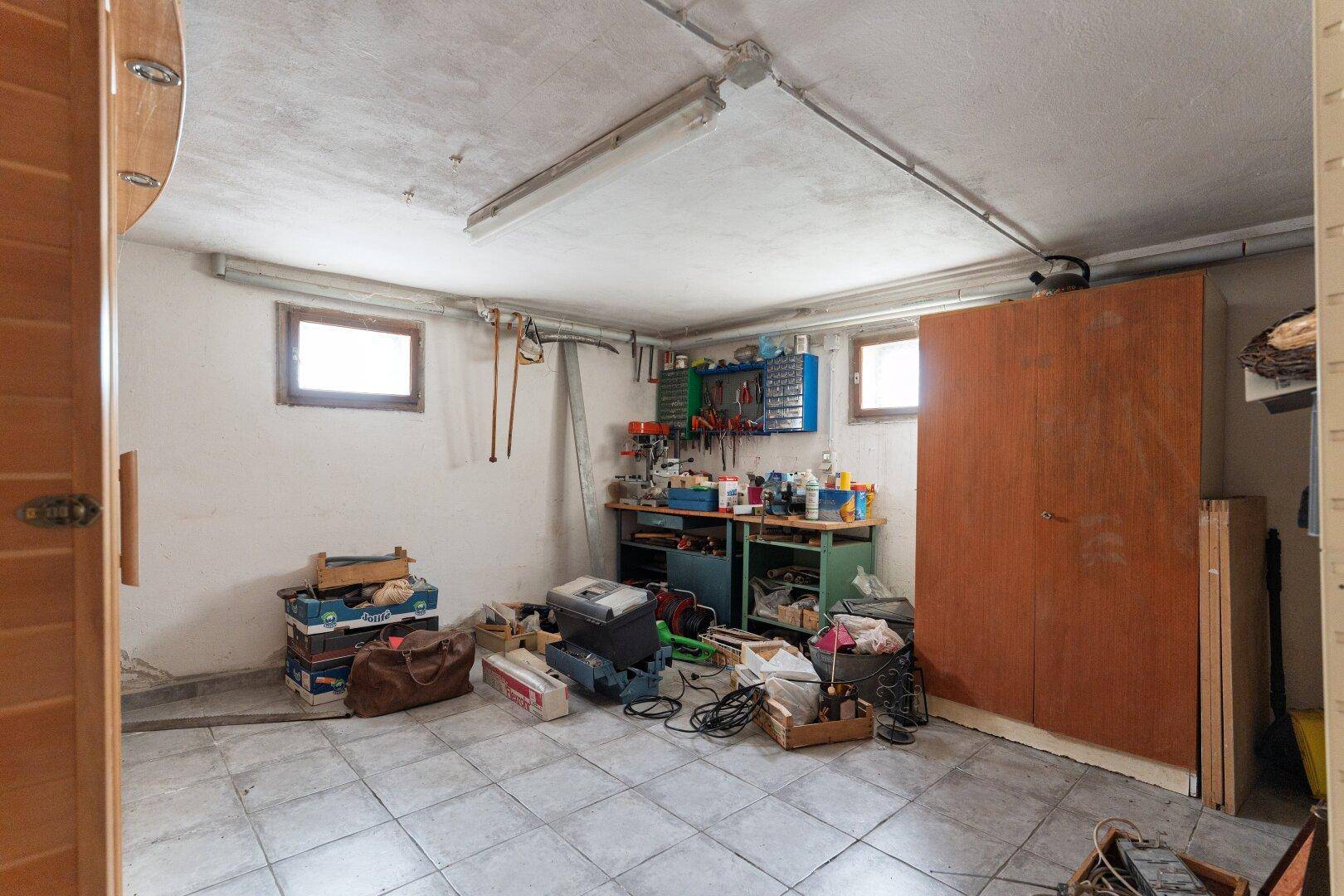 Kellerraum als Werkstatt und Lagerbereich mit Werkbank, Regalen und kleinen Fenstern.