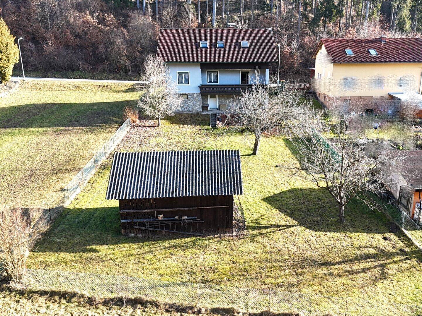 Weitläufige Ansicht des Grundstücks mit Haus, Nebengebäude und Waldrand.
