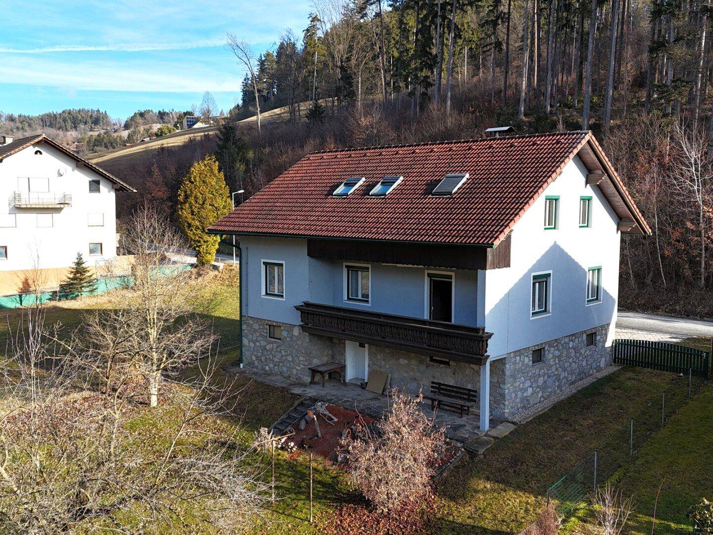 Frontansicht des Hauses mit Balkon und Steinsockel, eingebettet in grüne Landschaft.