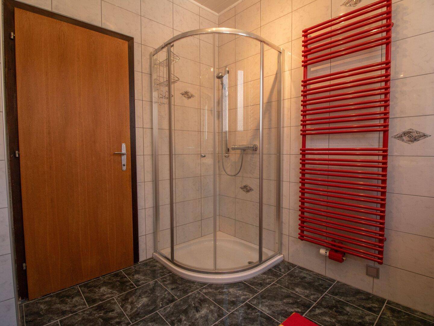 Badezimmer mit Duschkabine und rotem Handtuchheizkörper, gefliest in hellen Tönen.