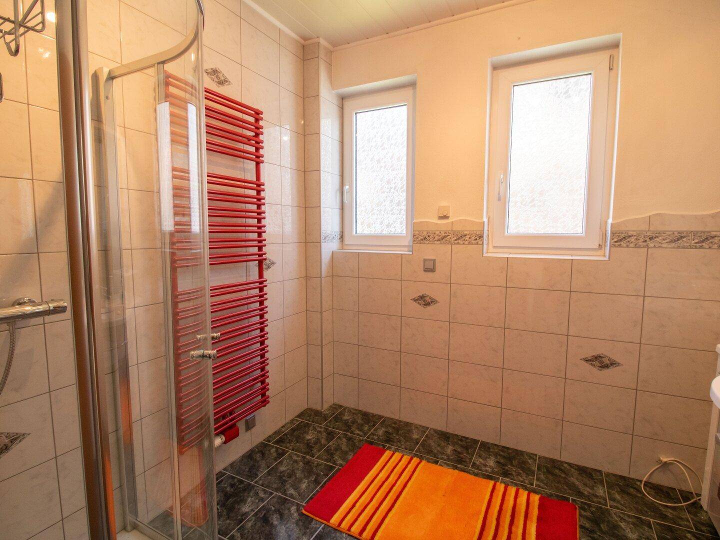 Helles Badezimmer mit Fenster, Duschkabine und rotem Handtuchheizkörper.