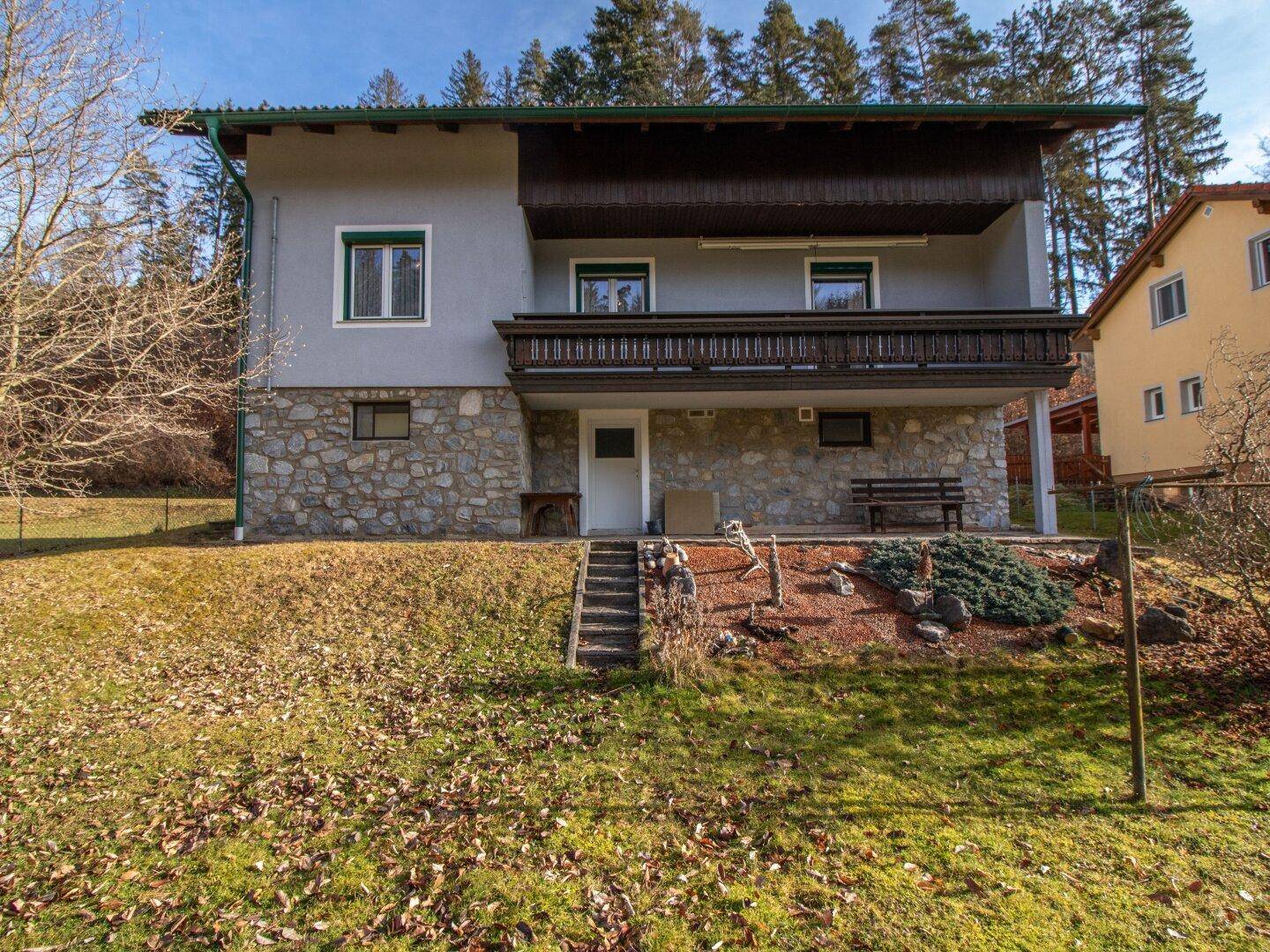 Rückansicht des Hauses mit Balkon, Steinsockel und kleiner Terrasse im Grünen.