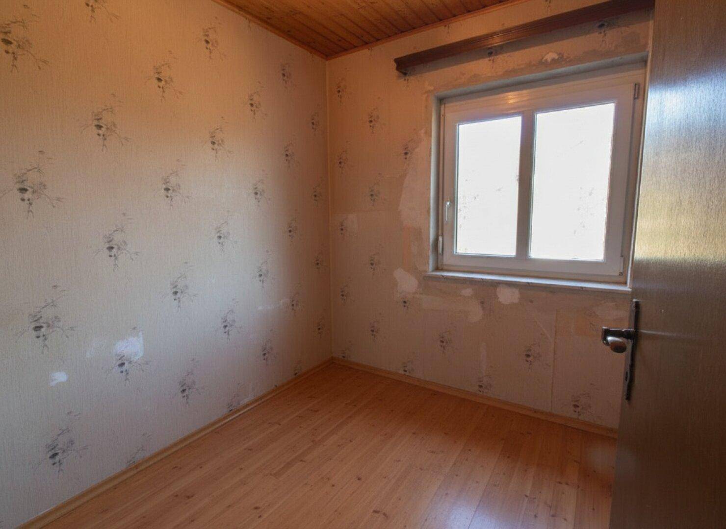 Schlafzimmer mit Holzboden und Fenster, Tapete muss erneuert werden.