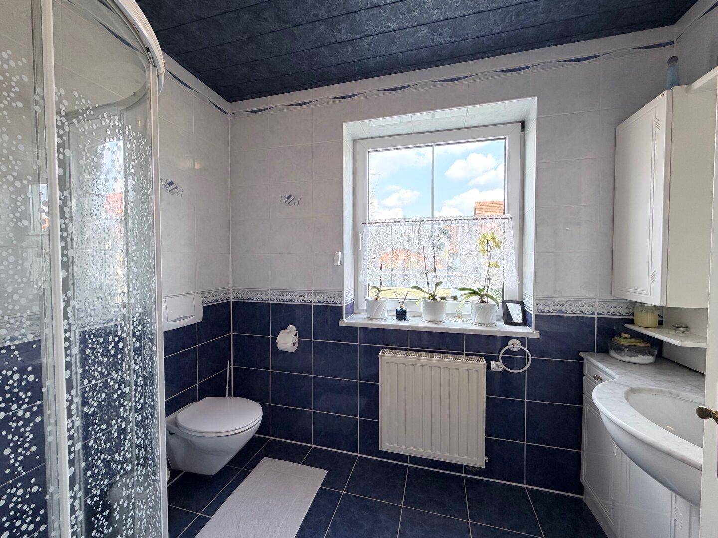 Badezimmer mit Dusche, WC, Fenster und blauen Wand- und Bodenfliesen.