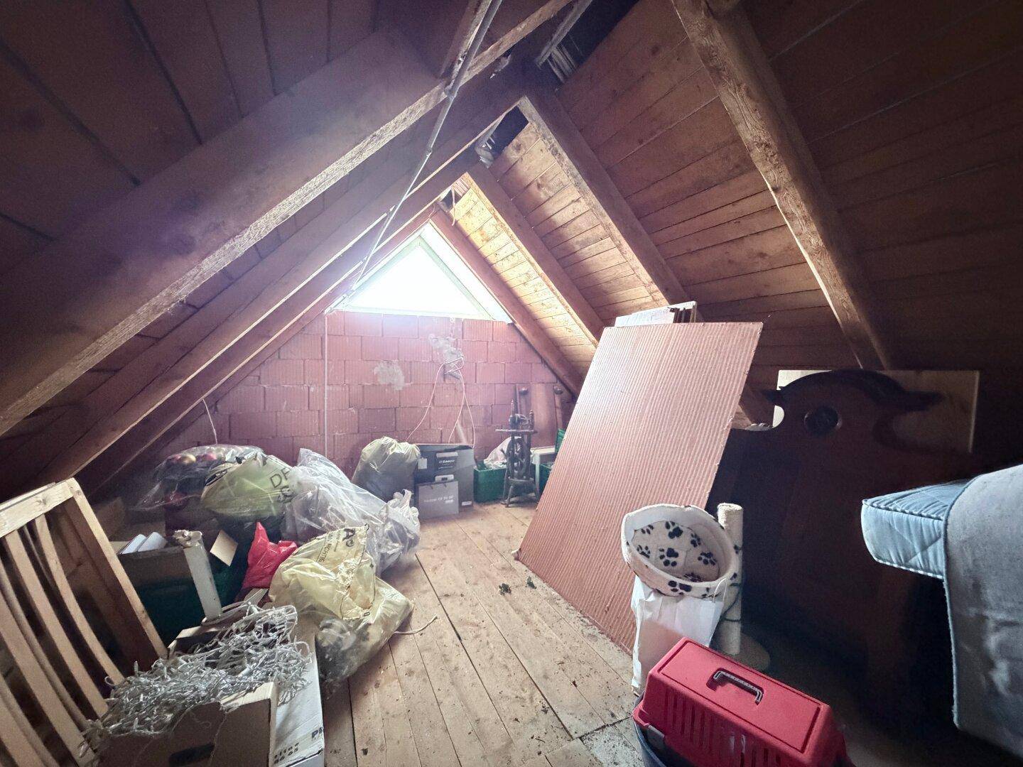 Dachboden mit sichtbaren Holzbalken und Stauraum, beleuchtet durch ein kleines Giebelfenster.