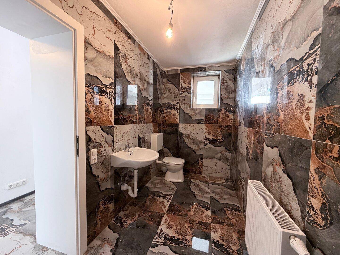 Badezimmer mit dunklen, gemusterten Fliesen an Wänden und Boden, ausgestattet mit Toilette und Waschbecken.