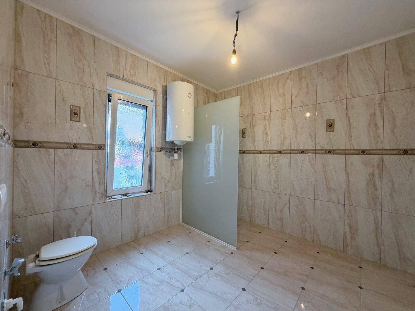 Badezimmer mit beigen Fliesen, Toilette und einer Duschabtrennung aus Glas.