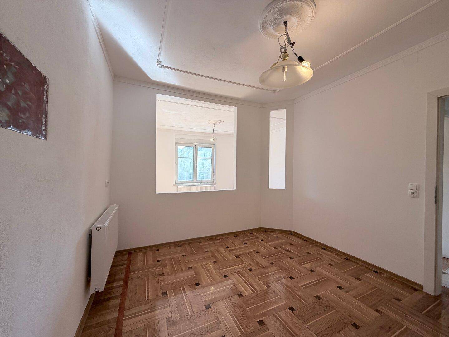 Heller Raum mit Parkettboden und einem Fenster, das Tageslicht hereinlässt.