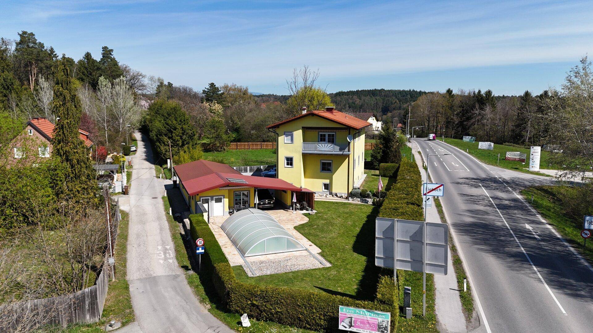 Luftaufnahme des Wohnhauses mit Garten, Poolüberdachung und angrenzender Straße.