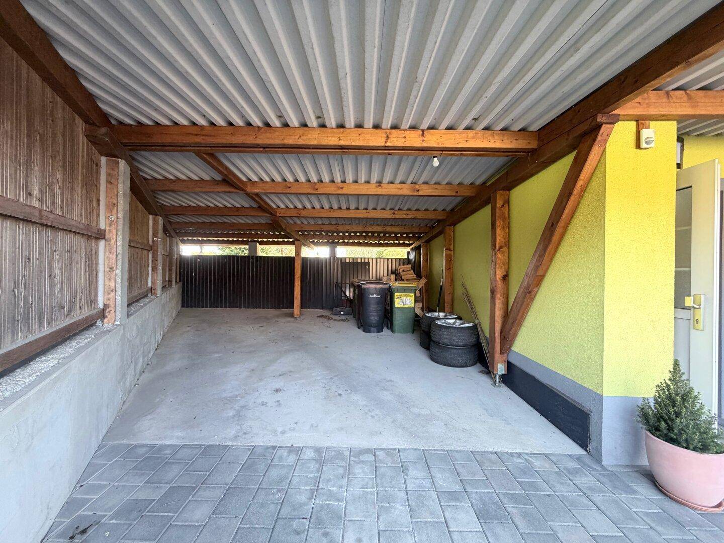 Überdachter Stellplatz mit Holzbalkenkonstruktion und gepflastertem Boden.