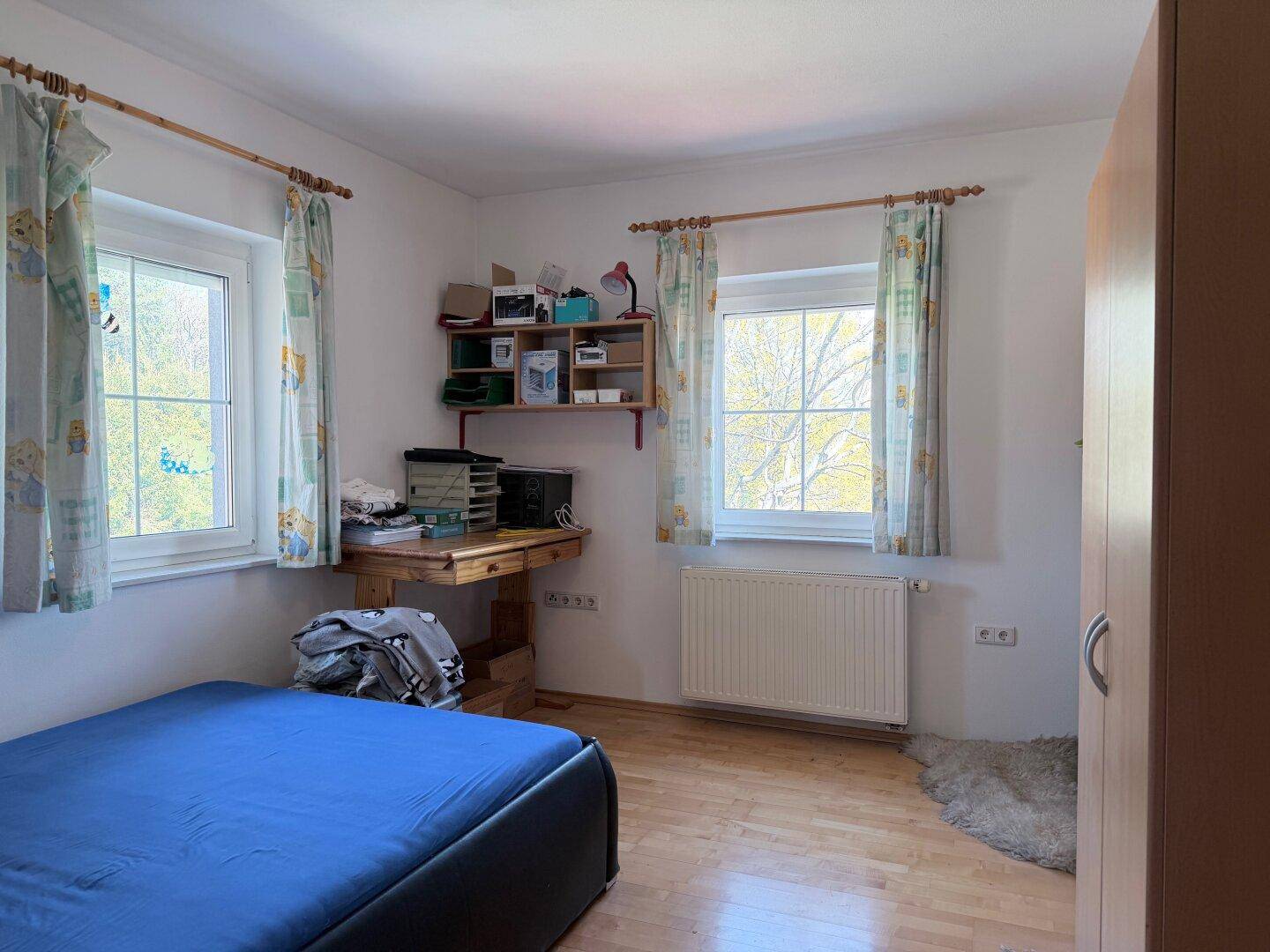 Schlafzimmer mit Holzboden, zwei Fenstern und einfacher Einrichtung.
