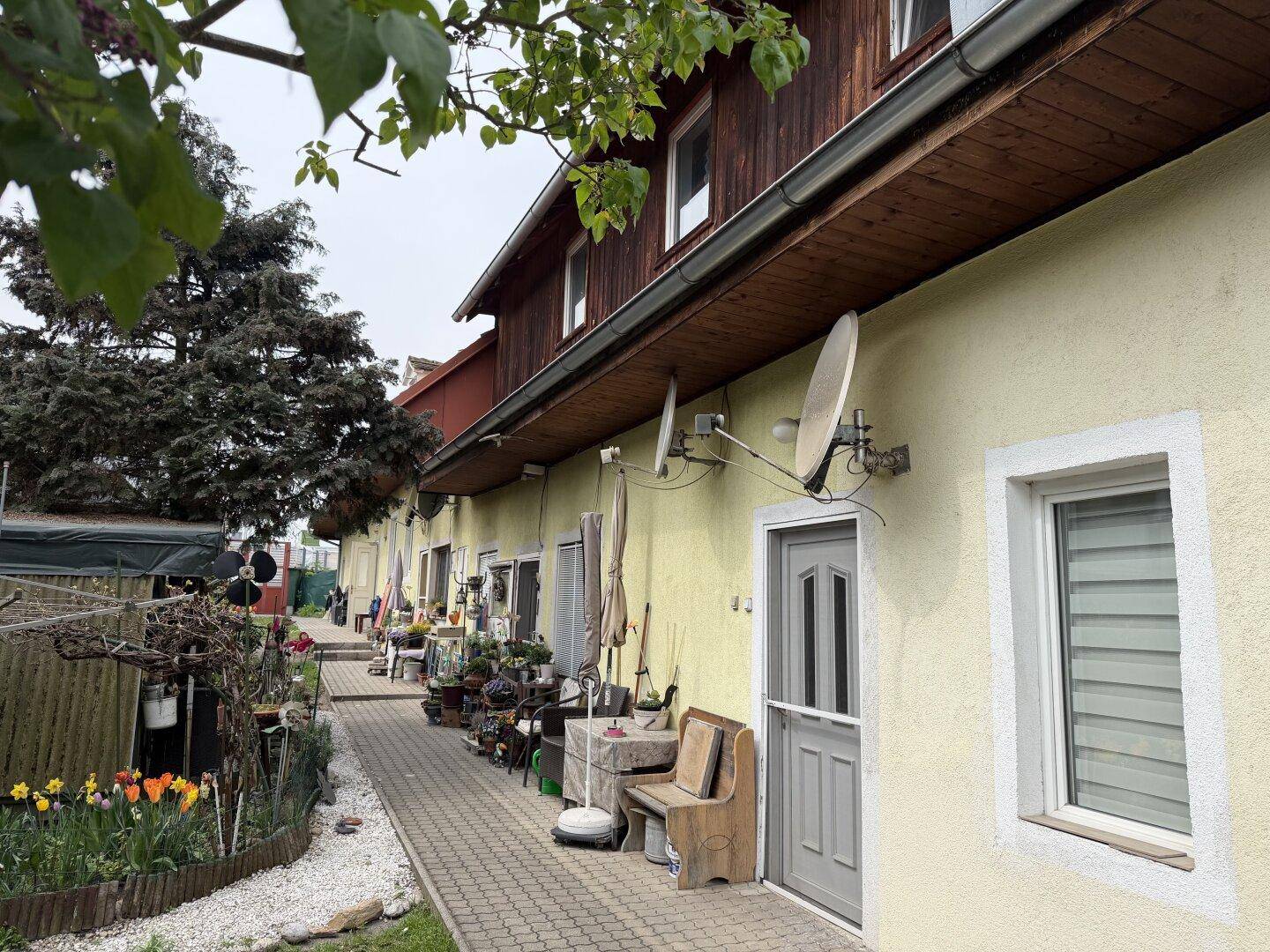 Gepflasterter Weg entlang der Hausfassade mit Blumenbeeten, Satellitenschüsseln und einem blühenden Baum.