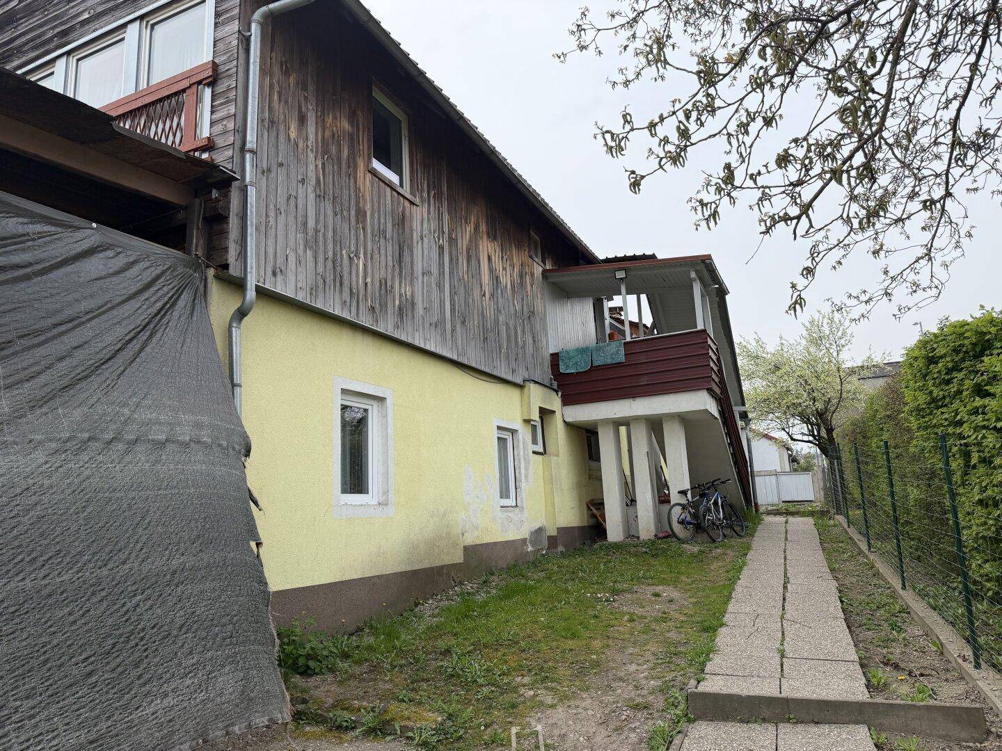 Seitenansicht des Hauses mit gelber Fassade, Holzverkleidung und einem kleinen Balkon mit Treppe.