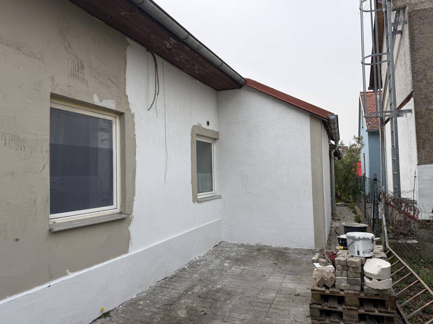 Hauswand in Renovierung mit teilweise neuem Putz und Farbe, daneben Baumaterialien und eine Leiter.