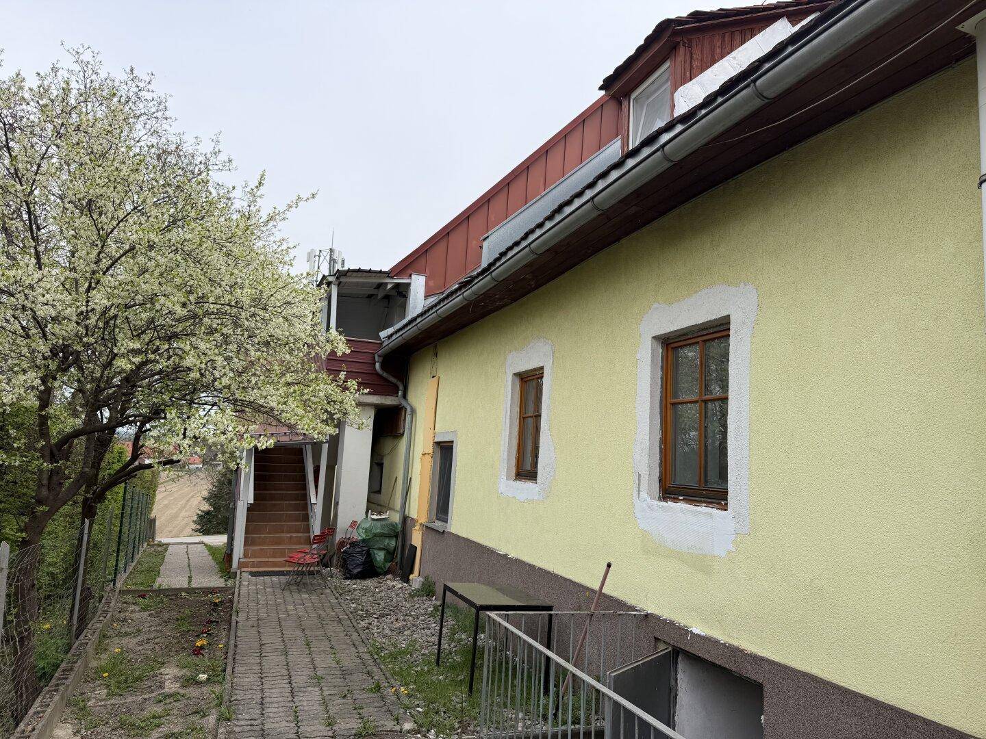 Seitenansicht des Gebäudes mit gelber Fassade, Holzfenstern und Außentreppe zum oberen Bereich.