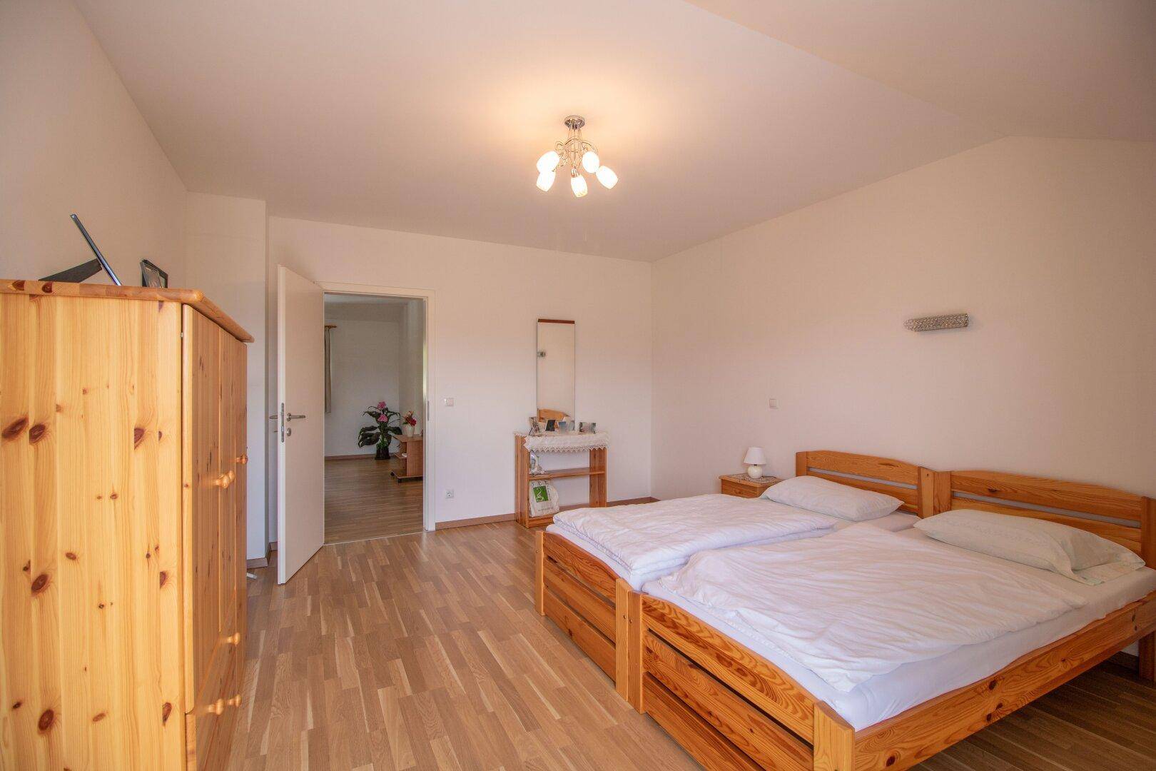 Helles Schlafzimmer mit Holzbett, Holzschrank und Zugang zu einem weiteren Raum.
