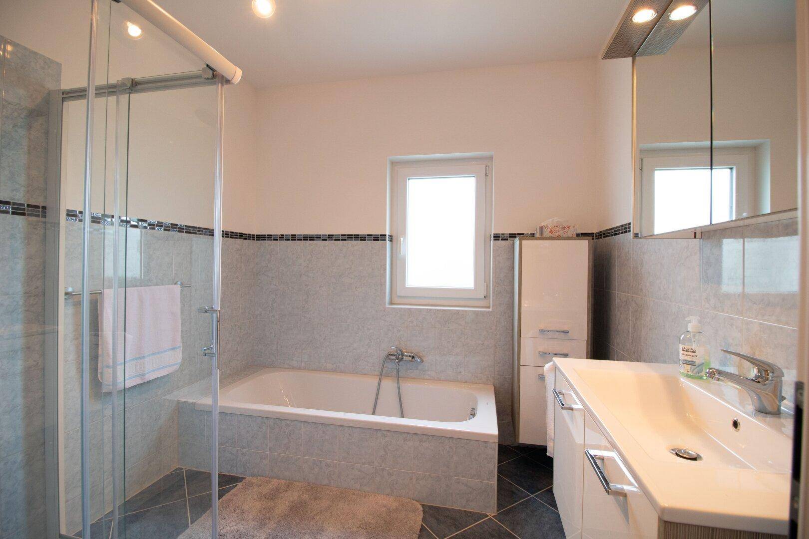 Badezimmer mit Badewanne, Duschkabine, Waschtisch und Fenster.